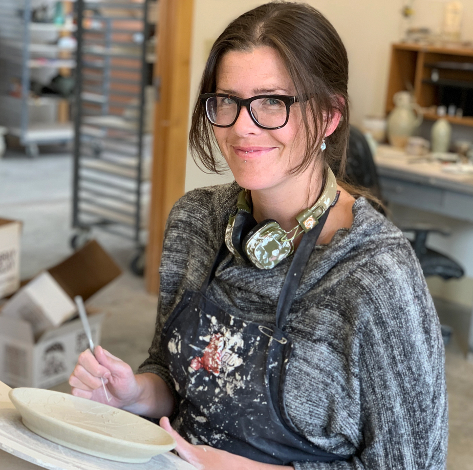 A women wearing glasses working on a piece of pottery in an arts studio.