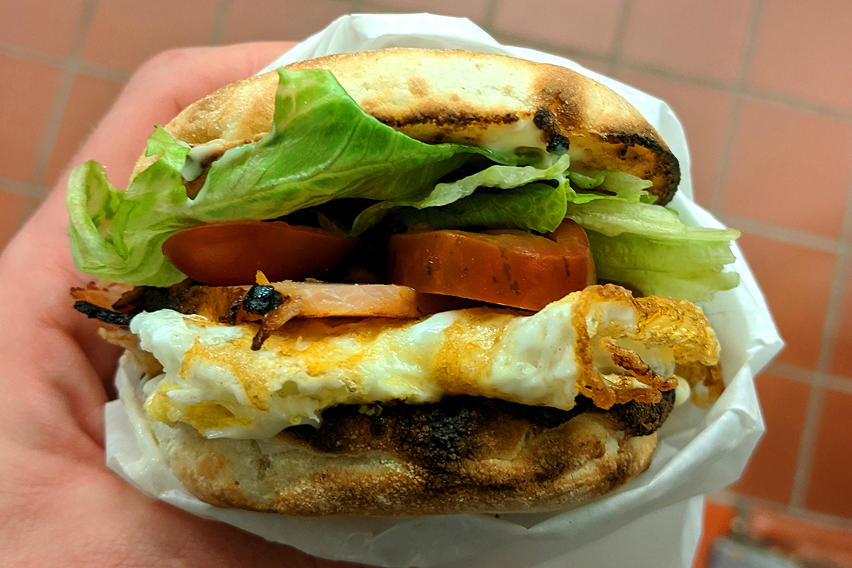 A hand holding a breakfast bun with egg, tomato and lettuce