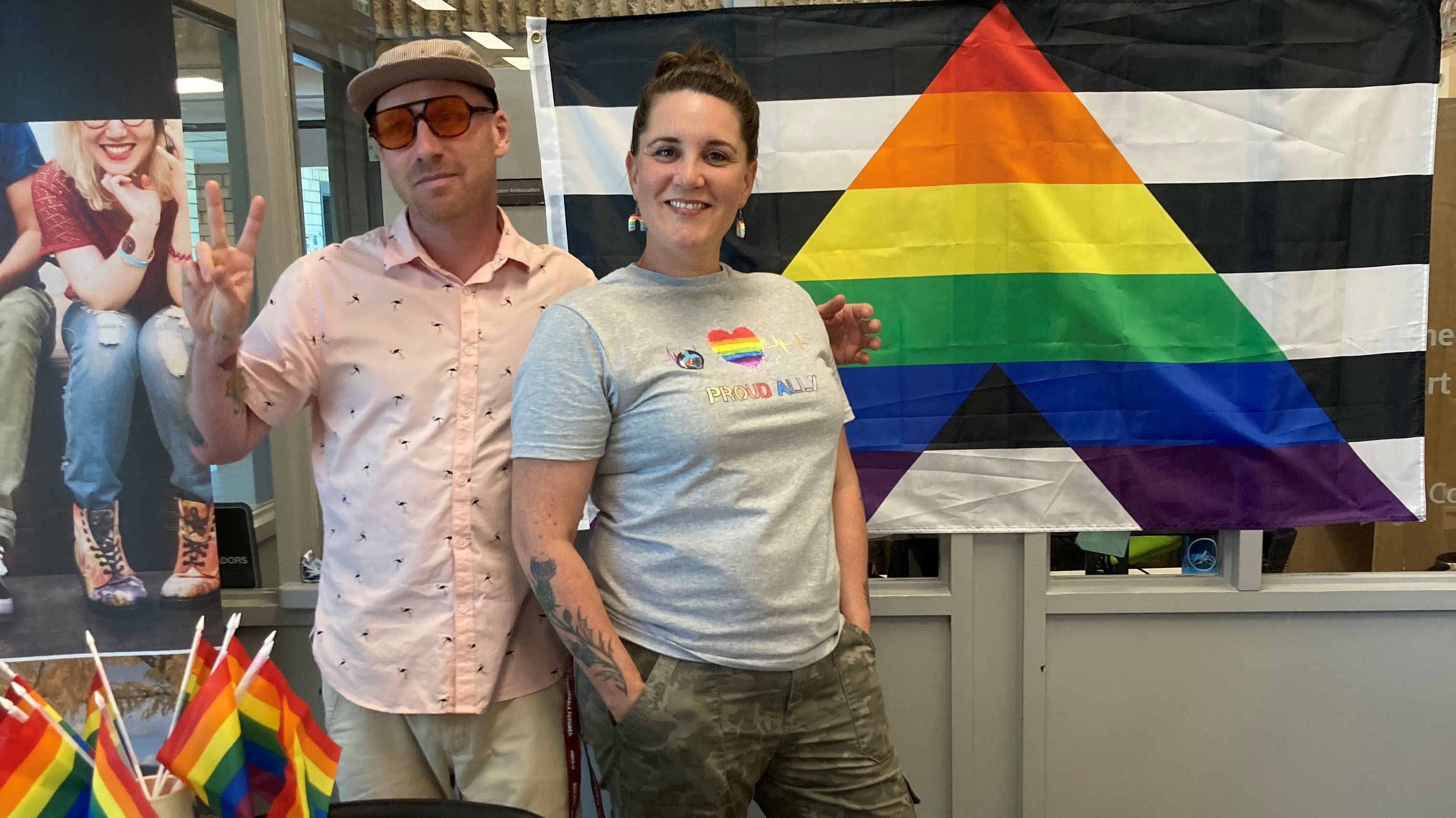 Two people stand smiling in front of a flag with a rainbow triangle
