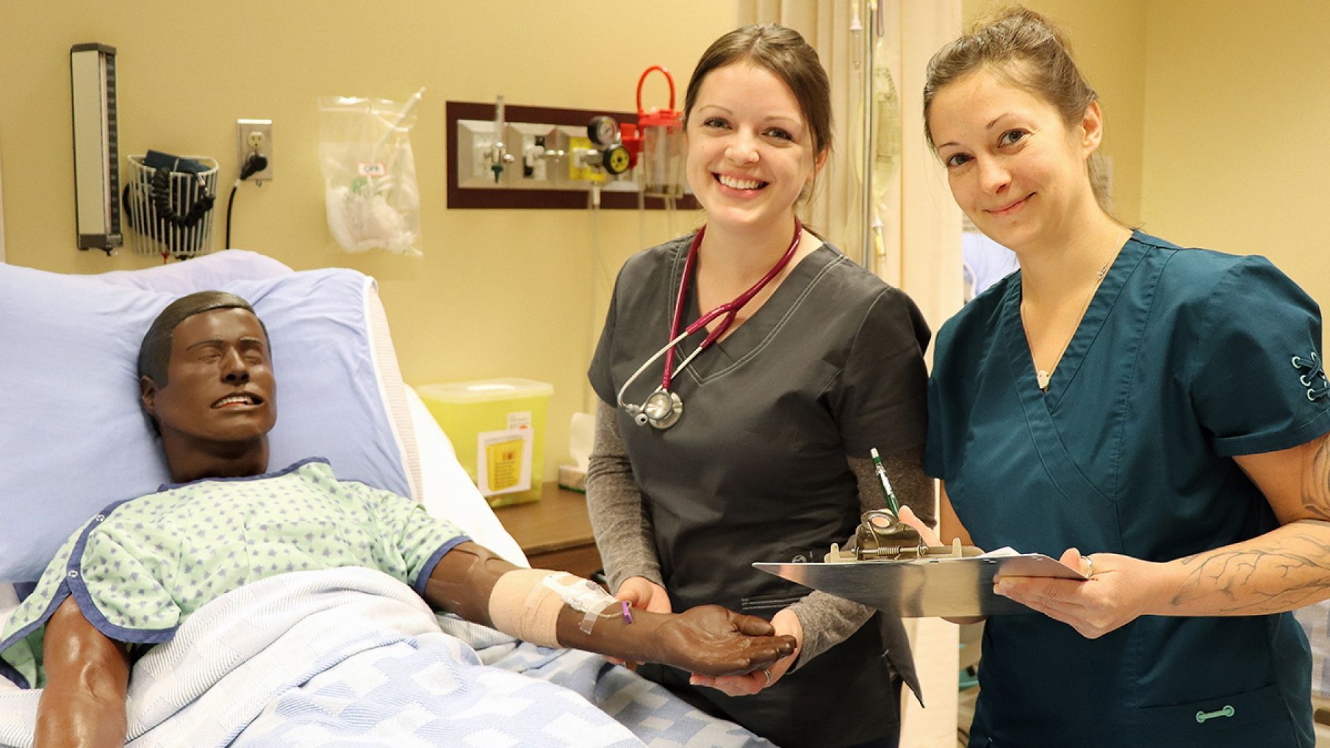 Two students stand in a hospital setting with a manikin