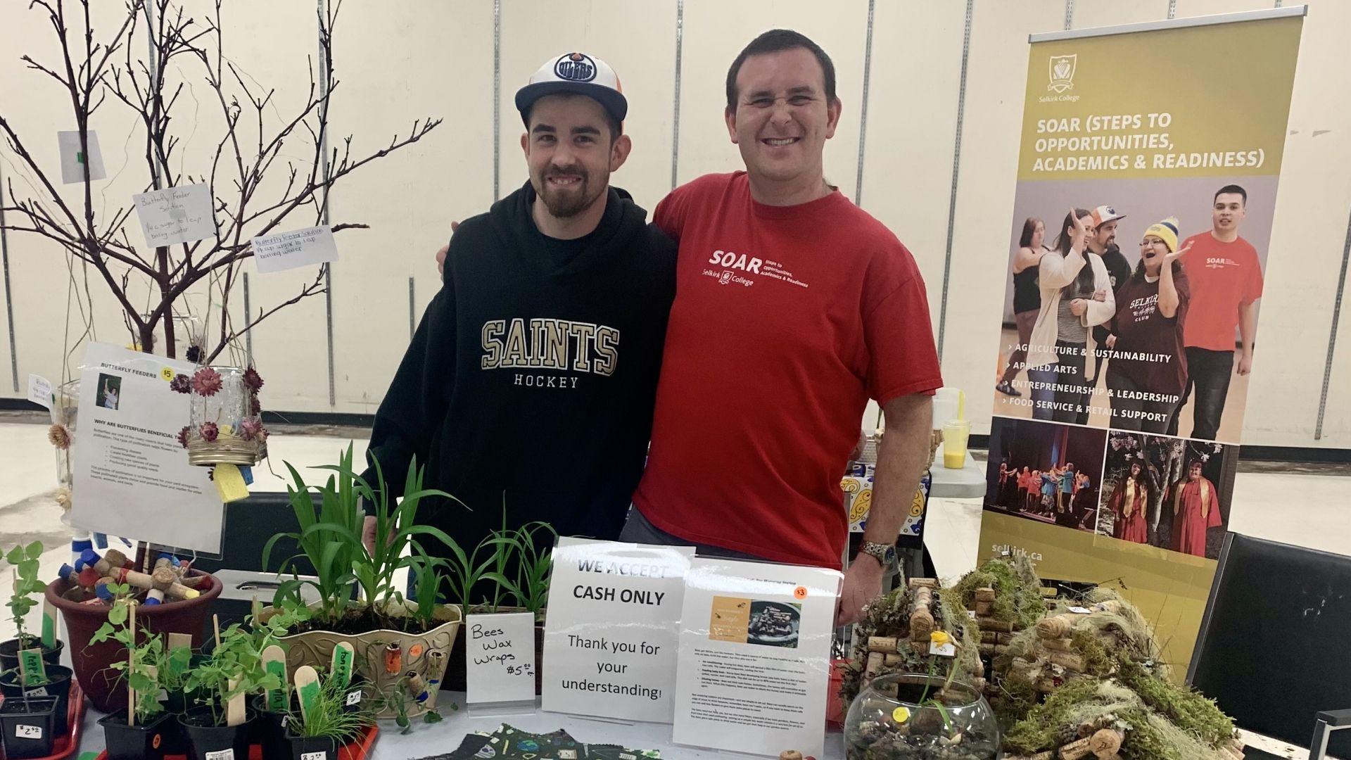 Two male Selkirk College students stand at a table selling plants and other hand-made items.