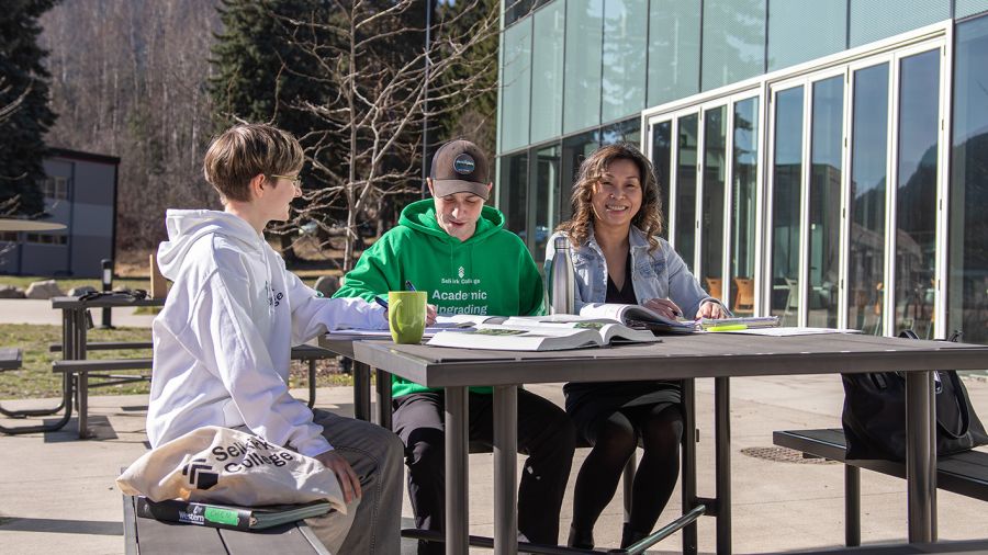 Three students sit outside at a picnic table with books
