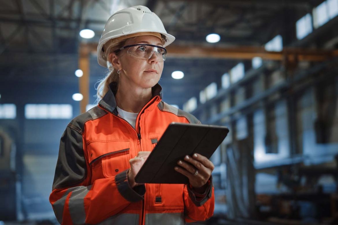 A woman wearing a hard hat stands in a factory and takes notes