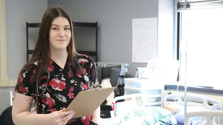 A Practical Nursing student stands in the lab holding a clipboard