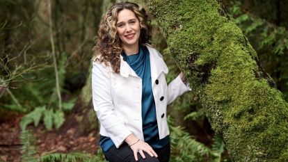 A woman stands in a forest next to a moss-covered tree