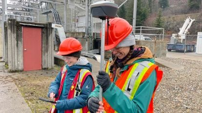 Two people wearing hard hats and high vis vests hold pieces of equipment
