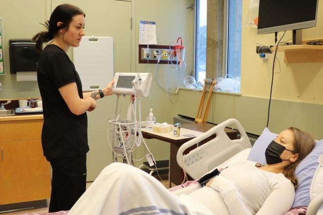 A nursing student stands at the bedside of a mock patient in a hospital bed