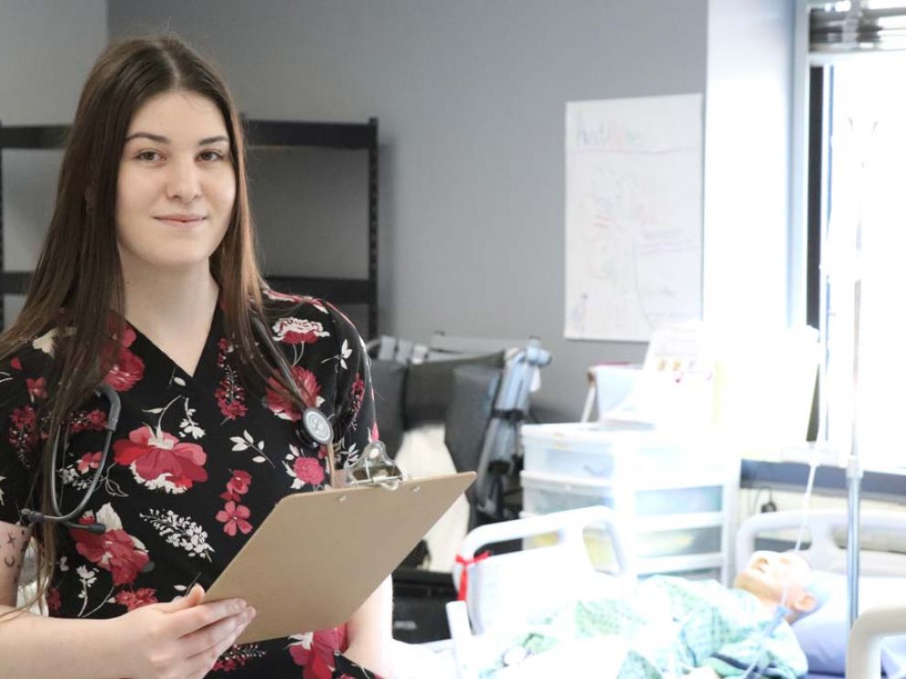 A Practical Nursing student stands in the lab holding a clipboard